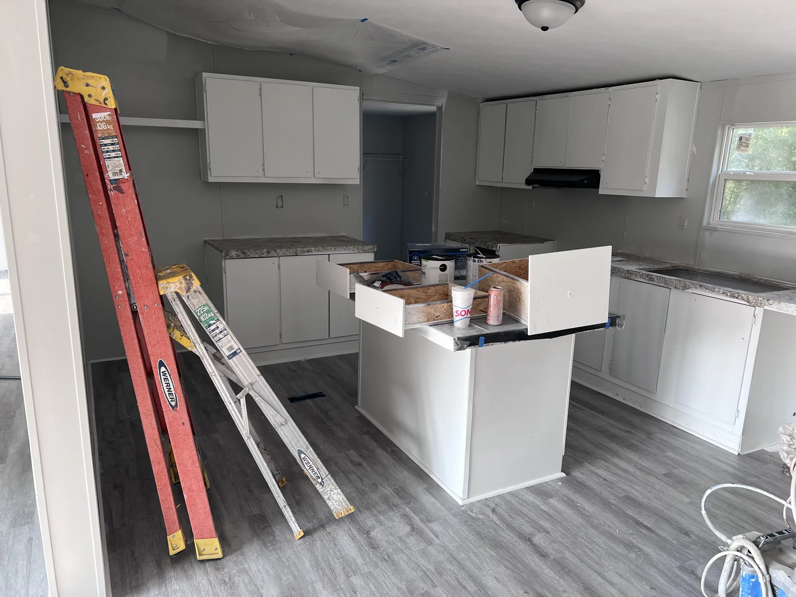 Kitchen during installation with white cabinets, an island, a ladder and grey flooring
